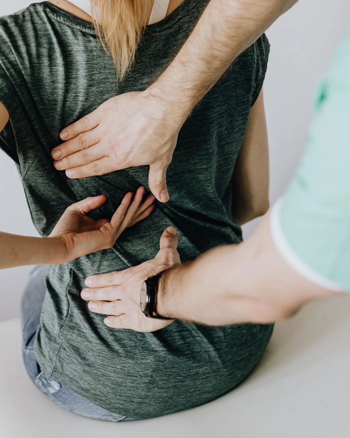 Une patiente assise, vue de dos, pose sa main sur le bas de son dos pour montrer la douleur ; les mains du praticien se posent au même endroit